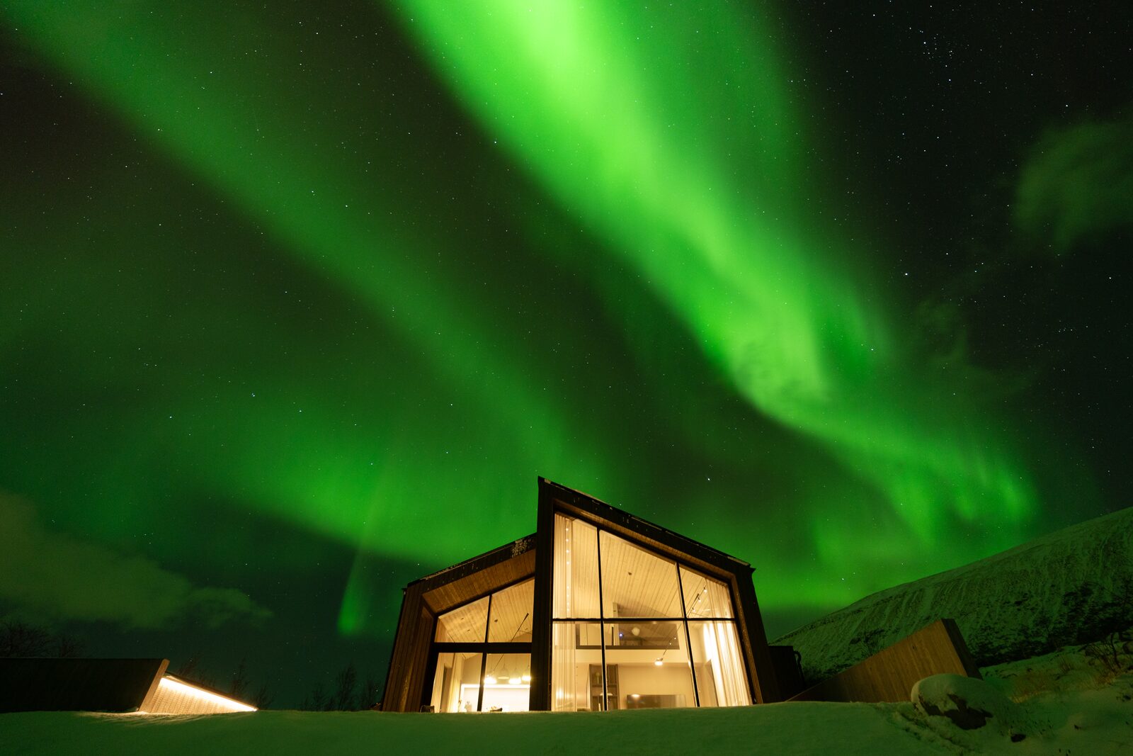 The house lit from within at night under a bright green aurora, with snow on the ground and hills behind.