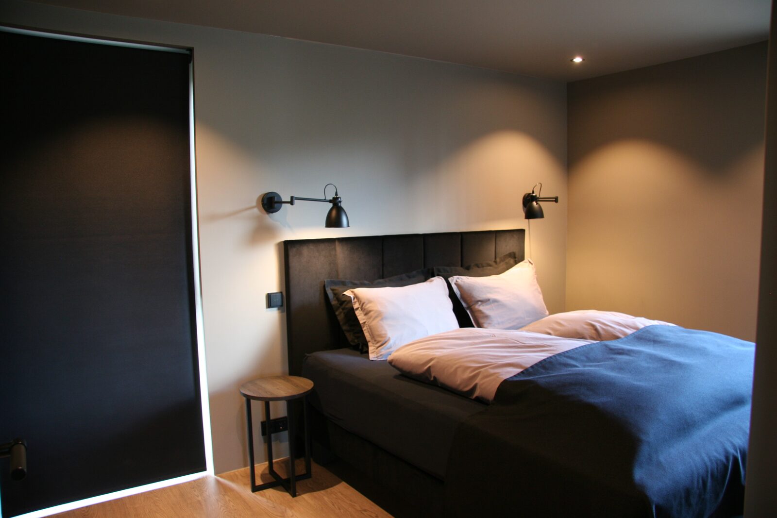 A queen-size bed with a dark headboard and wall sconces, daylight coming through a partly-lowered blackout blind.