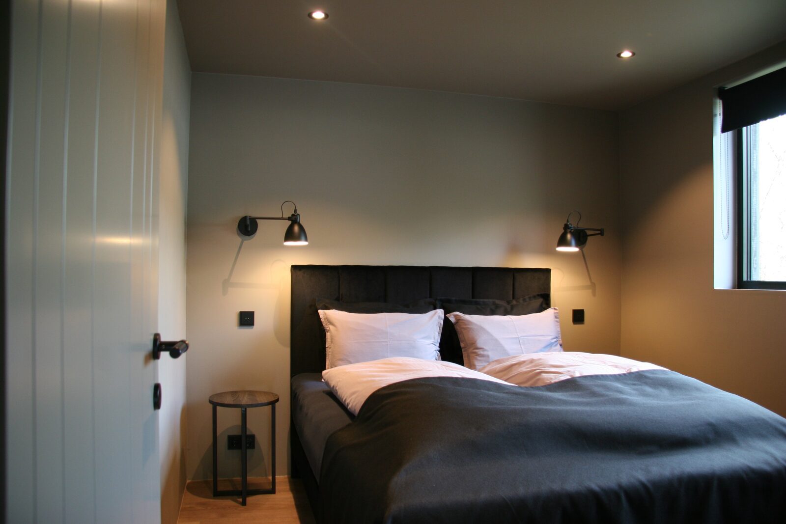 A queen-size bed with a dark upholstered headboard flanked by black articulated wall lamps, a round side table and a door to the left.