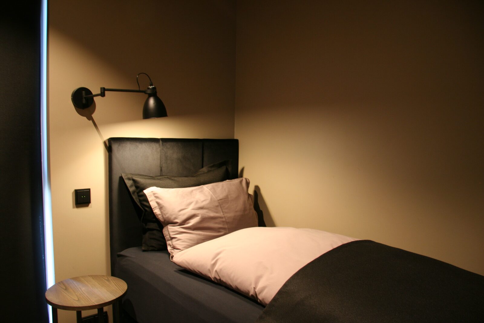 A single bed with a dark frame and pink-toned pillow beneath a black wall sconce.