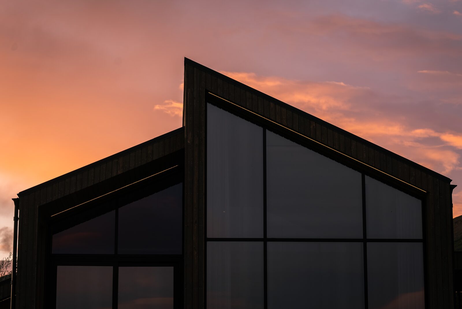 The asymmetric gable of the house in silhouette against a pink and orange sunset, the tall glass wall divided by two mullions and a transom.