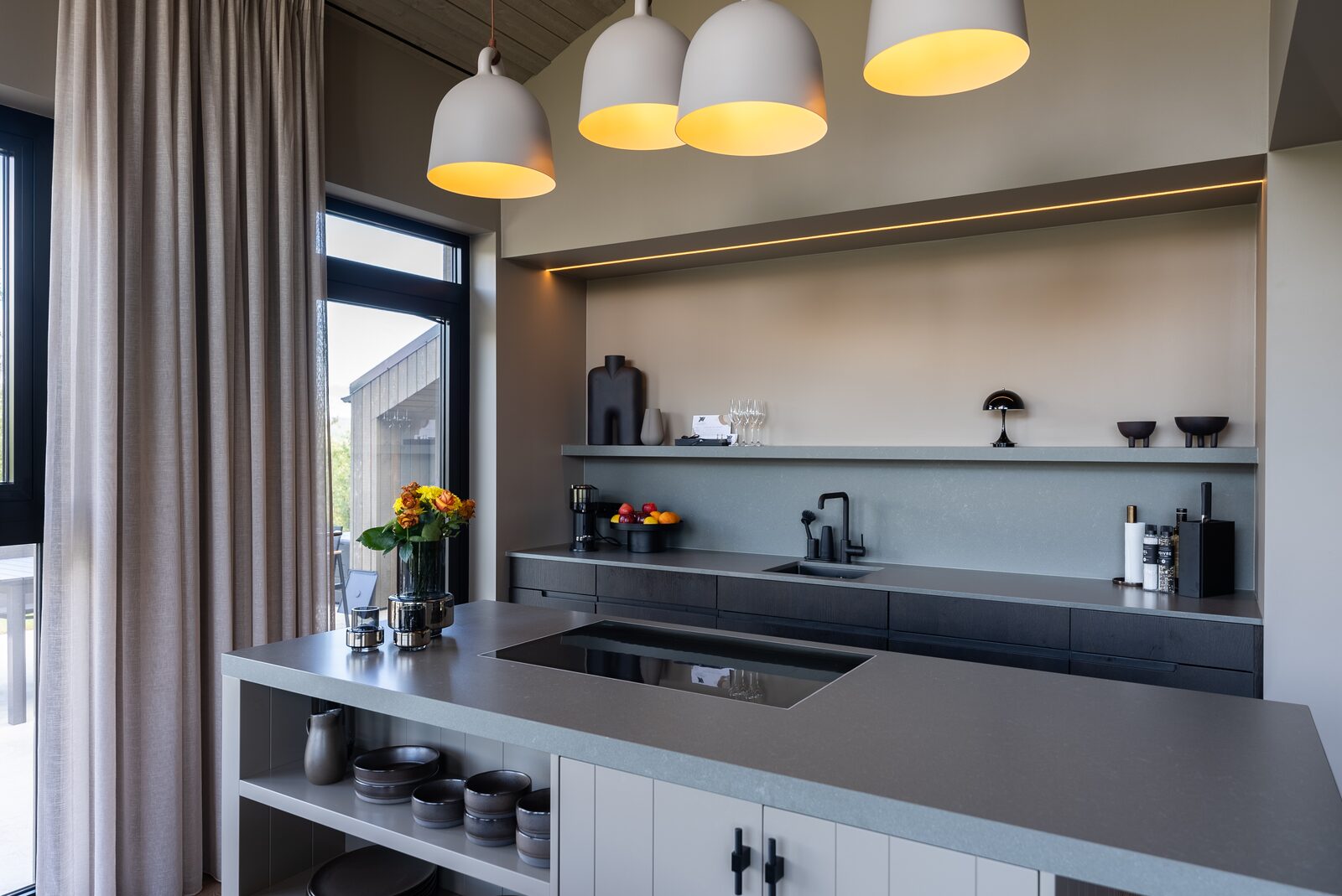 A dark kitchen counter beneath four dome pendant lights, with fresh flowers and a bowl of fruit.