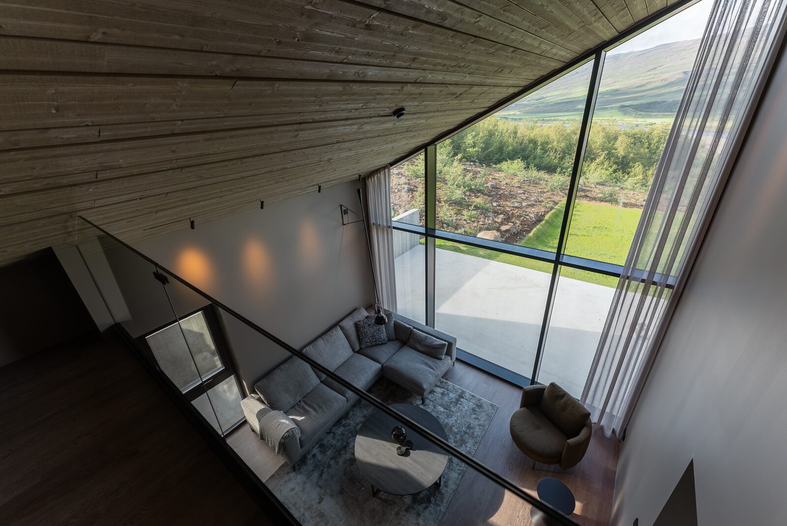 Looking down from the upstairs landing over the empty living room, sofa and coffee table below, full-height windows to the valley.