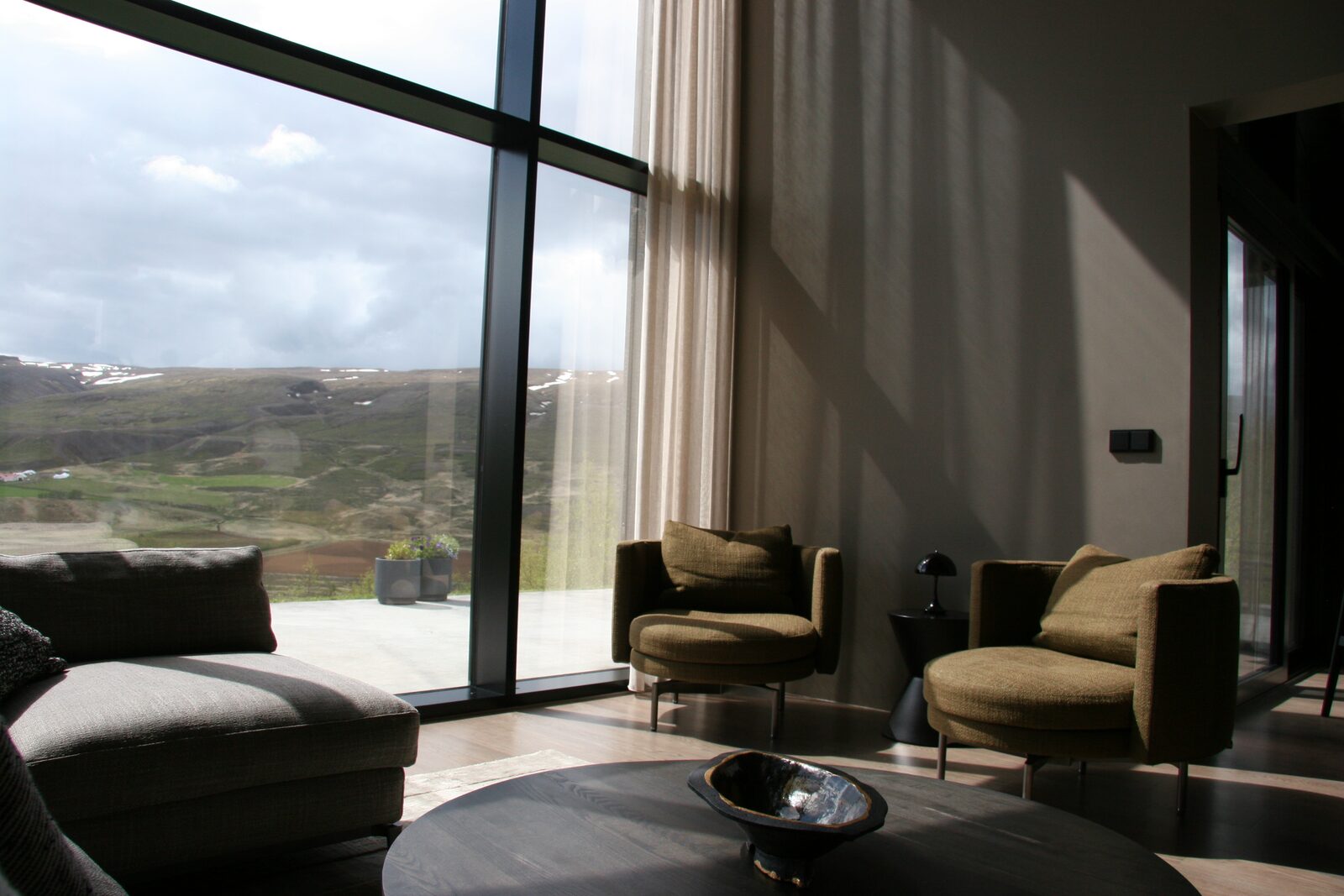 A grey sectional sofa and armchairs beside floor-to-ceiling windows looking out over the valley in daylight.