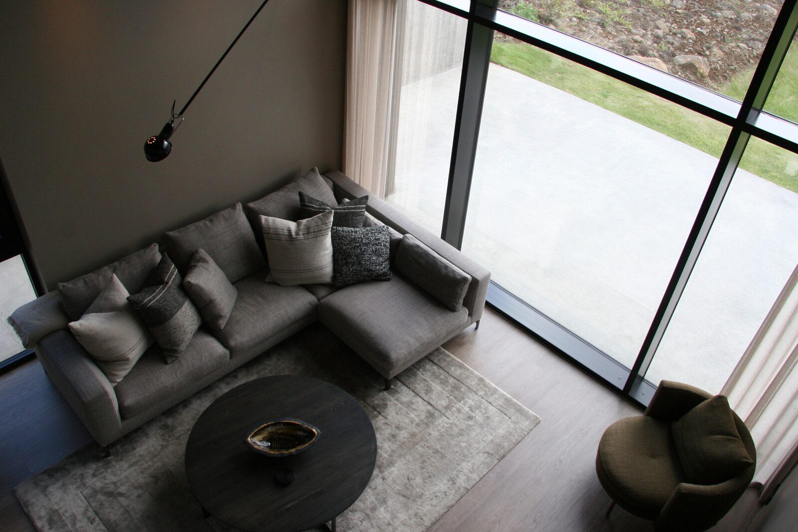 A grey sectional sofa and round coffee table with a floor-to-ceiling glass wall opening onto the paved terrace.