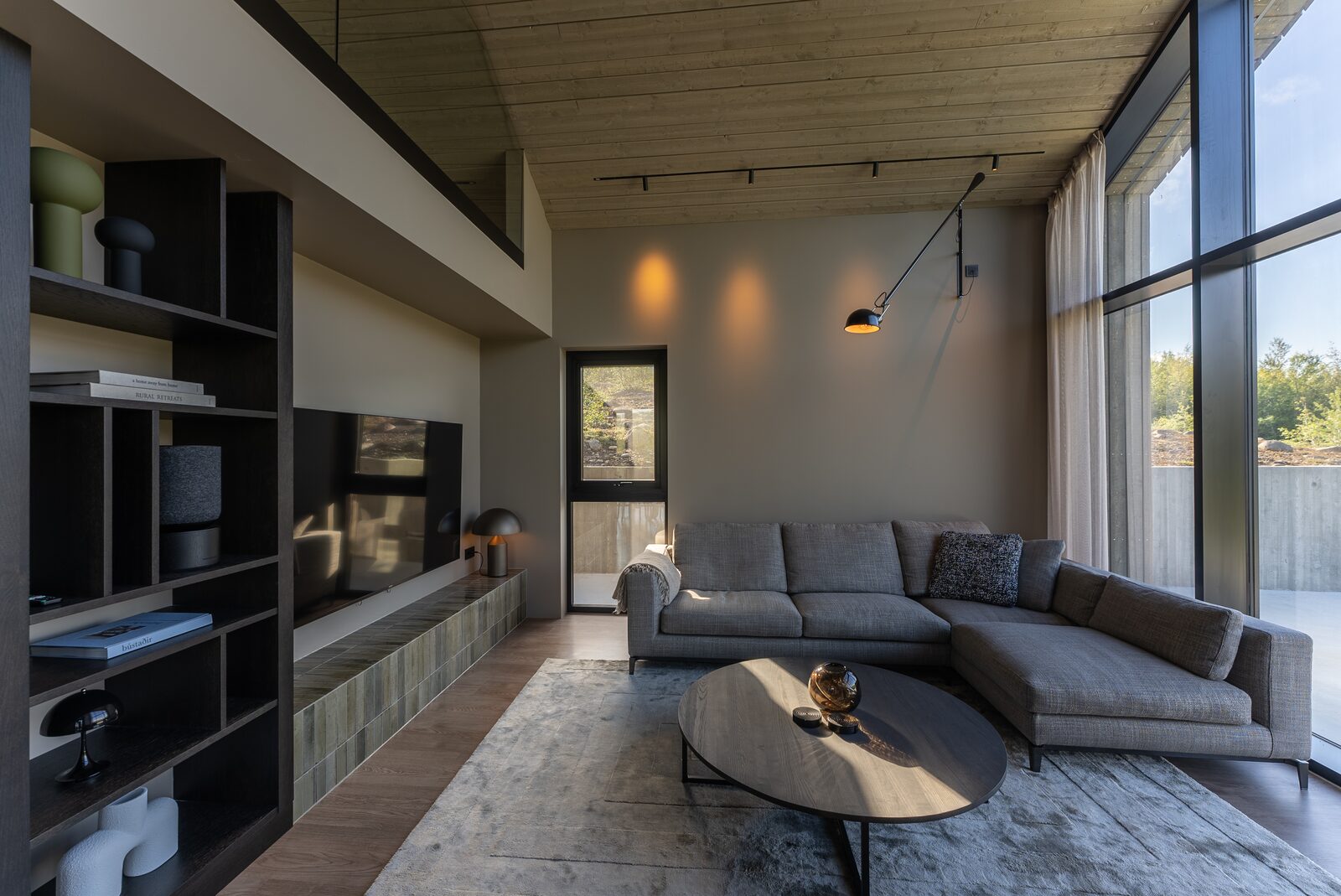 A wide view of the living room with a low sectional sofa, round coffee table and wall-mounted shelving unit, full-height windows to the view.