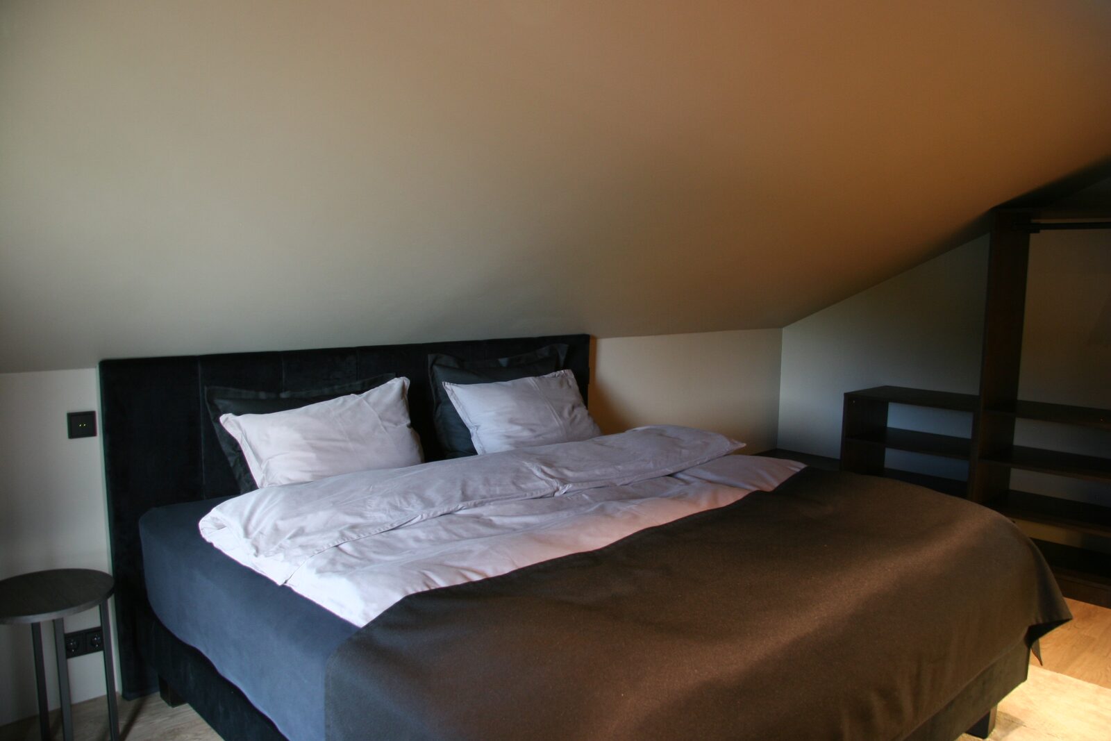 A king-size bed with a black upholstered headboard under a sloped ceiling, with open wardrobe shelving to one side.