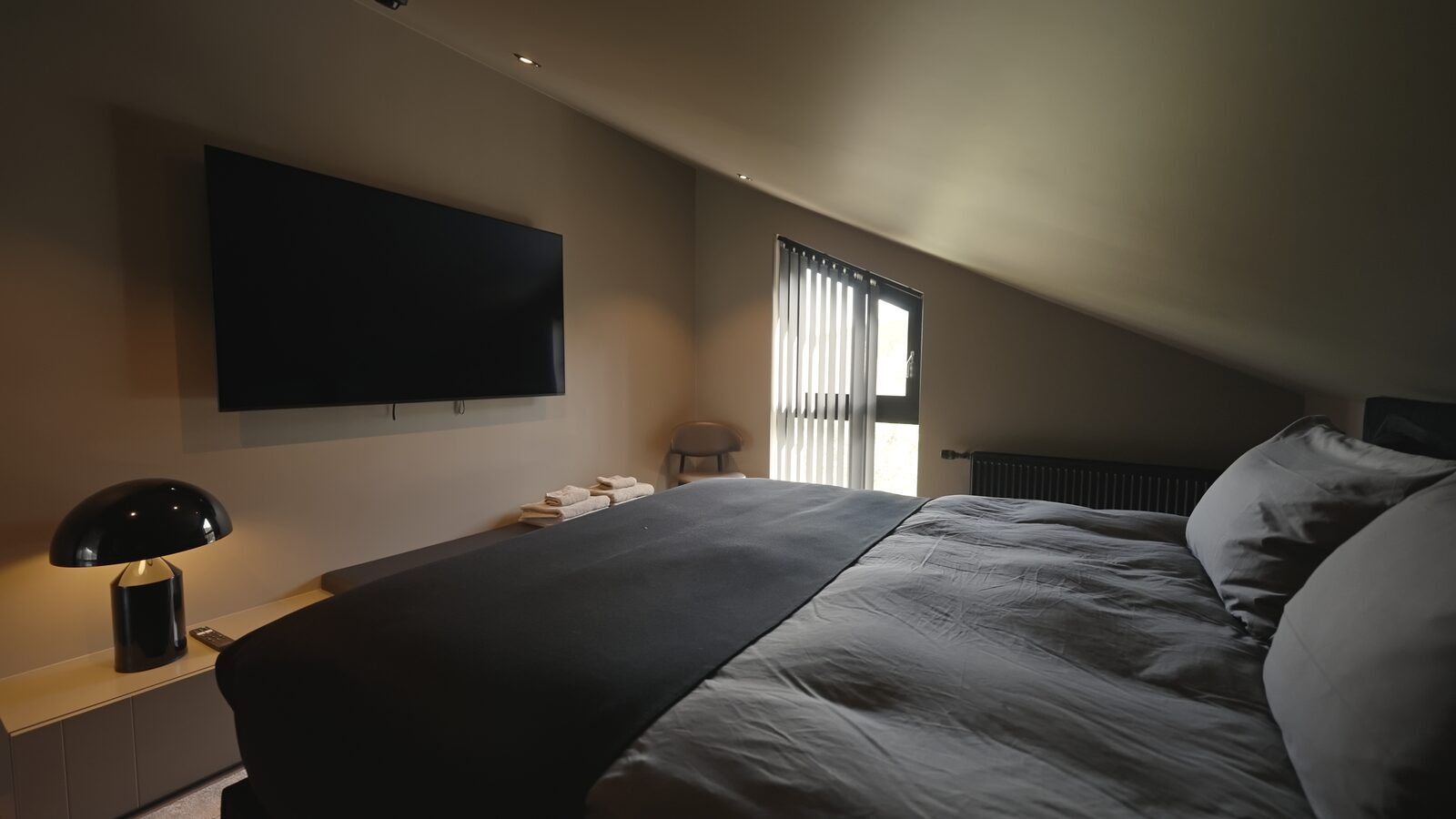 The master bedroom's sloped-ceiling wall with a mounted TV and a black mushroom-shaped lamp, the bed and a curtained window alongside.