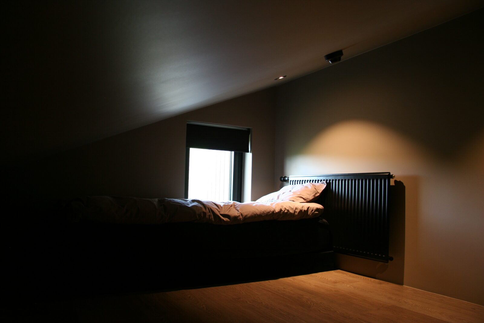 A single bed positioned beneath a window, next to a dark panel radiator, in dim evening light.