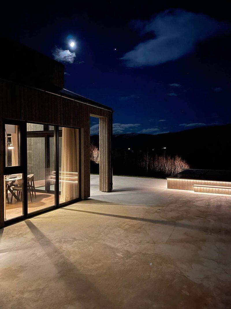 The terrace at night under a bright moon, with light from the dining room, the lit hot tub at the edge and bare birches beyond.